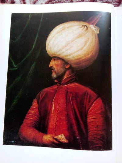 The image shows a painting of a man in profile, facing left. He is wearing a large, cream-colored turban and a vibrant red robe with gold trim. The background is dark and features flowing green drapery. The man has a beard and appears to be holding a small, light-colored object in his right hand. The painting is framed by a white border and the corner of a textured, maroon and white patterned surface is visible at the top of the image. There is no discernible information within the image that would indicate a location in the city of Zhmerynka, Ukraine.