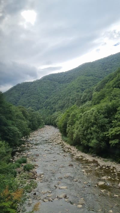 A wide-angle, vertical, high-angle shot shows a rocky riverbed with shallow, clear water flowing downstream between densely forested, green mountains. The sky is overcast with patches of bright white light breaking through the clouds. The mountains are covered in a lush, green canopy of trees. The river is shallow, with numerous rocks and pebbles visible both in the water and along its banks. The overall impression is one of a natural, wild landscape.