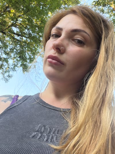 The image is a close-up, low-angle, selfie-style shot of a woman outdoors, likely during the daytime. She has blonde hair and is wearing a grey ribbed tank top. A purple strap is visible over her left shoulder, and a tattoo can be seen on her left arm. The background consists of a bright blue sky and the green leaves of a tree. The woman is looking slightly off-camera with a neutral expression. There is no discernible connection to the city of Telgte, Germany in the image.