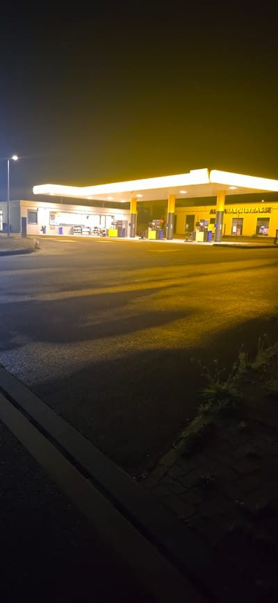 The image captures an outdoor scene at night, featuring a brightly lit gas station. The gas station has a large, yellow, illuminated canopy supported by yellow and grey columns. Beneath the canopy, multiple gas pumps are visible, some appearing blue and yellow. To the left of the pumps, there is a building with windows and a sign that reads "AUTO-WASCHSTRASSE". The ground in front of the gas station is asphalt, with visible shadows cast by the illuminated structures. The sky is dark, with some ambient light contributing to the yellow and white tones of the scene. The overall impression is that of a typical, operational gas station at night.
The specific location is the AUTO-WASCHSTRASSE (Car Wash Street) in Seevetal, Germany.
