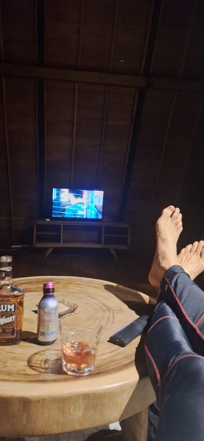 The image depicts a domestic scene within a wooden room, possibly a loft or attic due to the angled ceiling constructed of wooden planks. In the background, a flat-screen television is mounted on a wooden entertainment unit. The television displays a person, and text below reads "Padel Training TV."

In the foreground, a large, round wooden table occupies the center of the frame. On the table, there are two bottles, one labeled "RUM Whisky," and a small bottle with a purple cap. A glass filled with an amber liquid, presumably whiskey or rum, sits in front of these bottles. To the right of the glass, a black remote control is placed on the table.

The lower right portion of the image shows a person's legs and feet, indicating they are relaxing and facing the television. The person is wearing dark pants with red trim. The feet are bare.

The overall impression is one of a relaxed evening spent indoors, with entertainment and drinks. The location is specified as Banjar, Indonesia, suggesting this scene is from a residence or lodging within that city.