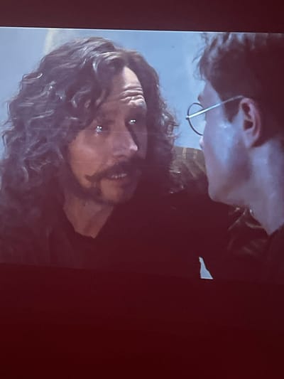The media file is a close-up, vertically oriented photograph of two individuals. The primary subject, in the foreground and taking up most of the frame, is a person with dark, curly hair. Their face is tilted upwards and to the left, with their eyes open and looking directly at the camera. They have a mustache and a beard. Two small, circular stud earrings are visible in their earlobes. The lighting suggests a dark setting, possibly indoors or at night, with a dramatic spotlight illuminating the face and hair.
Behind and slightly above this main subject is another individual, whose face is largely obscured. Only their upper face, including an ear and part of their forehead, is visible. This person is wearing thin, silver-rimmed glasses that curve around their ear. Their skin appears lighter in tone than the main subject. The background is dark and out of focus, with a subtle suggestion of a lighter, possibly textured surface in the upper left corner. A small, irregular white shape is also visible in the upper right quadrant of the image against the dark background. The overall mood conveyed by the image is intense and intimate due to the close proximity of the subjects and the direct gaze of the primary individual. There is no visible text in the image. The location context of Gliwice, Poland, does not offer any specific visual cues within the image itself.