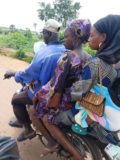 The image shows three people riding on a motorcycle in Iseyin, Nigeria. The person in the front is a man wearing a blue jacket and a cap with "APC" visible on it. He is wearing glasses and appears to be driving. Behind him, a woman is seated, wearing a colorful patterned dress and a headscarf. She has large hoop earrings. The third person, also a woman, is seated behind the first woman and is wearing a black headscarf and a patterned dress with blue and white polka dots. She has gold hoop earrings and is carrying a woven gold purse. Both women appear to be wearing several layers of clothing, including a folded blanket or cloth in bright colors. One of the women has a small green shoe with a Nike logo on it. The scene appears to be a rural or semi-rural area with greenery and trees visible in the background. The ground is dirt. The lighting suggests it is daytime, likely overcast given the diffused light. There is a small sign with "100 PACE" visible on the motorcycle's rear fender.