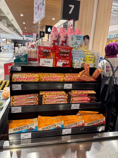 The image provides a detailed view of a product display in a supermarket or grocery store in Kelapa Dua, Indonesia. The scene is brightly lit, indicating an indoor, daytime setting.

**Subjects:** A person, likely an employee, is partially visible from the back, wearing a purple head covering (hijab) and a light-colored apron. A cardboard cutout featuring a smiling young man, labeled "Napas Dulu," is placed among the products.

**Objects:** The central focus is a black retail display stand filled with various confectionery items. On the left, there are "Take-it" chocolate bars, including "Ovaltine DELFI TAKE IT OVAL" (13,500 Rp) and "4 FINGERS" (11,800 Rp). Above them are multiple packs of "Ricola" Swiss Alpine Herbs candies, with varieties like "LEMONMINT" priced at 22,500 Rp and other flavors at 22,000 Rp.

The main sections of the display showcase "SilverQueen" chocolate bars, specifically "Milk Chocolate with Cashews" (15,800 Rp) and "CHUNKYS" variants (28,000 Rp). On the right, "VAN HOUTEN" chocolate bars are prominent, featuring "dark milk chocolate," "43% Cocoa," and "dark milk almonds" options, all priced at 16,500 Rp.

Other visible objects include checkout lane numbers "5", "6", and "7", a "PROMOTION EXPRESS" sign, and a "KILLS TOILET BACTERIA" cleaning product with "www.cleanshop.co.id" on its label. A partial "AKERY" sign (likely part of 'BAKERY') is visible in the background.

**Activity:** The products are neatly arranged for sale, suggesting a typical retail shopping environment where customers select items, possibly near a checkout counter.

**Notable details:** All prices are clearly displayed in Indonesian Rupiah (Rp). The visible text provides extensive brand and product information, including "BE BOLD MORE COCOA" and "SINCE 1828" for Van Houten. Social media handles like "@Ponsto" are partially visible on the person's uniform.