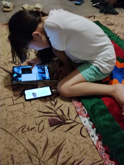 In Almaluu, Kyrgyzstan, a young person with dark hair, wearing a white t-shirt and turquoise shorts, is seen lying on a carpeted floor. The person is engaged with two electronic devices: a tablet and a smartphone. The tablet is open and displaying a screen with what appears to be a video or game interface. The smartphone, placed below the tablet, also shows a lit screen with some content visible. The surroundings include other footwear and a colorful blanket or rug. The lighting suggests an indoor environment, possibly a home.