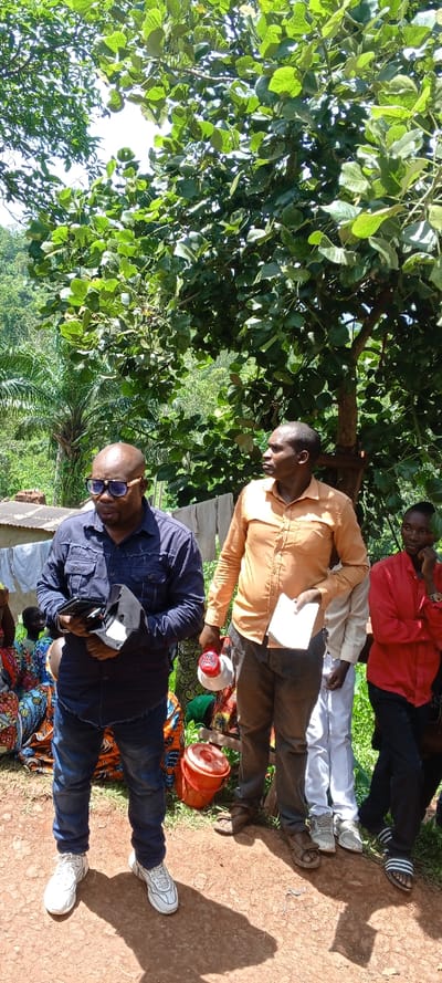 In a rural setting, four men stand on reddish-brown soil near trees and foliage. The man on the left wears a blue denim shirt, jeans, white sneakers, and sunglasses. Next to him, another man in an orange shirt and grey pants holds a megaphone and papers. Partially visible, a man in a white shirt and the last man in a red shirt are behind them. In the background, people are seated on the ground, and there's a building with clothes hanging outside.
