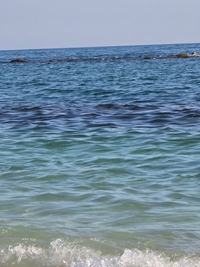 The image is a vertical, eye-level shot of the sea with gentle waves. The water transitions in color from a deep blue at the horizon to a lighter, more translucent turquoise near the foreground. In the distance, a line of dark rocks, partially submerged, breaks the surface of the water, with a small patch of green vegetation visible on one of them. The sky above is a pale, clear blue. The scene is devoid of any human presence or man-made structures, suggesting a natural coastal environment. Without further information, it is not possible to determine if this specific location is within the city of Salerno, Italy.