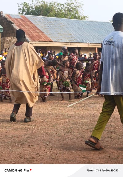 The image depicts a cultural scene in Gidan Kogi, Nigeria. In the foreground, two adult figures frame the shot, one wearing a beige robe and dark trousers, and the other in a grey "Planning Committee" t-shirt and green trousers. In the mid-ground, a group of children dressed in patterned clothing with red and brown tones are performing a dance or ritual. Some hold small flags and green foliage. Behind them, a crowd of spectators and a building with a corrugated iron roof are visible. The ground is a reddish-brown color, suggesting earth or dirt.
