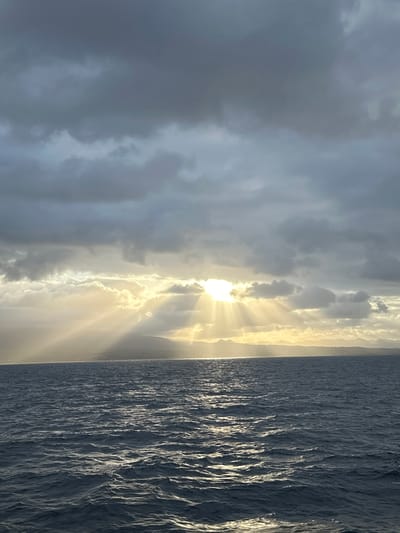 The image depicts a seascape with a dramatic sky. The sun is partially obscured by dark, heavy clouds, but its rays break through in a powerful display, creating a crepuscular effect over a distant, mountainous coastline. The sea in the foreground is dark blue and choppy, with light glinting off the wave crests. The overall impression is one of natural beauty and atmospheric intensity. The location is not identifiable as Capesterre-Belle-Eau, Guadeloupe from the visual information present.