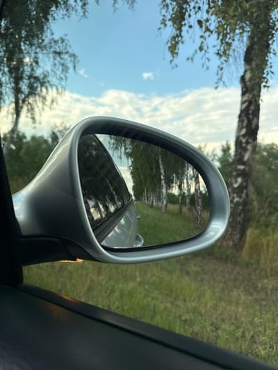 The image is a close-up shot of a car's side mirror, reflecting a rural landscape. The mirror, with its silver casing, shows a reflection of a road lined with trees and grass under a cloudy sky. The sky is a mix of blue and white clouds, suggesting a partly sunny day. Trees are visible on both sides of the road in the reflection, some appearing as tall, slender trunks with sparse foliage, possibly birch trees, while others are more densely leaved. The road itself appears to be unpaved, with hints of greenery along its edges. The perspective is from inside the car, looking out at the mirror. The scene is likely taking place during the daytime, given the lighting conditions and the visible sky. The overall impression is one of a serene drive through a natural environment. No people are visible, and no text is present in the image or its reflection. The presence of a car mirror suggests the context of travel. Based on the provided location context of Kalynivka, Ukraine, this scene depicts a typical rural view from a car in that region.