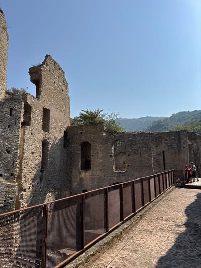 The image captures a view of the Castello dei Doria in Dolceacqua, Italy, a historic medieval castle. The perspective is from a pathway overlooking the ruins. The castle walls, constructed of rough stone, are visibly weathered and partially collapsed, showcasing arched window openings and crumbling battlements. A rusty metal fence with a mesh inlay runs along the pathway, providing a barrier. The pathway itself is paved with uneven stones and is partially cast in shadow. In the background, a lush, green hillside with trees rises against a clear blue sky. Two individuals are visible on a raised platform to the right of the frame, observing the castle.