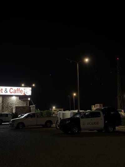 The image depicts an outdoor scene at night, likely in the city of El Gouna, Egypt. A lit sign for "t & Caffe" is visible on the left side of the frame. Several vehicles are present, including a white pickup truck and a black and white police vehicle with "POLICE" written on its side. A few individuals are standing near the white pickup truck. In the background, a tall communication tower with red lights is visible, along with some buildings and streetlights. The ground appears to be asphalt. The overall atmosphere suggests a nighttime commercial or public area.