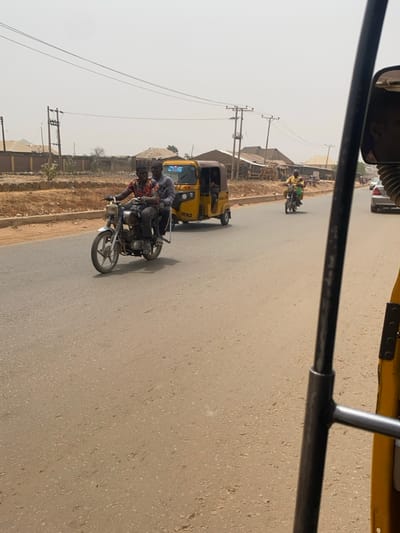 A daylight scene captured from within a yellow auto-rickshaw (Keke Napep) depicts a multi-lane asphalt road in a Nigerian urban or semi-urban environment. The road surface is dry, grey, and shows signs of use.
In the foreground to mid-ground on the left, two adult males are riding a silver-colored motorcycle. The operator wears a multi-patterned shirt (red, black, white), dark trousers, and a dark cap. The passenger, seated behind, wears a grey long-sleeved shirt, dark trousers, and a dark cap. They are traveling away from the camera.
Adjacent to the motorcycle, a yellow three-wheeled auto-rickshaw (Keke Napep), displaying "PANA" on its side, is in motion. At least two occupants are visible within its shaded passenger compartment. Further to the right and slightly behind, a third adult male, wearing a light yellow/orange long-sleeved top, operates a dark-colored motorcycle. A white sedan is partially visible in the far right background.
The left side of the road is bordered by a reddish-brown dirt shoulder. Beyond this, several single-story and multi-story buildings with light-colored walls and dark roofs are visible. Multiple utility poles with overhead wires are spaced along both sides of the road, extending into the hazy, light grey sky. Structures with corrugated metal roofs are also discernible in the background. The ambient light suggests bright, possibly dusty, conditions.