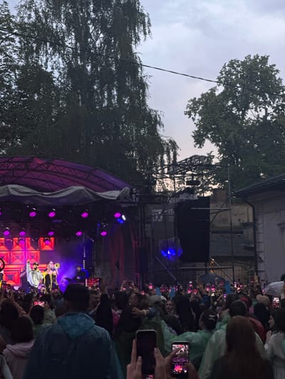 The image depicts an outdoor concert event occurring in Ivano-Frankivsk, Ukraine, with a crowd of people gathered to watch a performance on a stage. The stage is illuminated with pink and blue lights, and several performers are visible, including a person playing a trumpet and another at a keyboard. Many audience members are holding up smartphones to record the performance. The setting appears to be a public space, possibly a park or square, with trees and buildings visible in the background. The sky is overcast.
