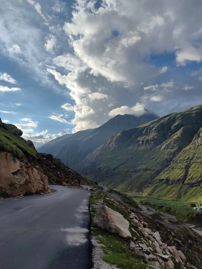 The image depicts a winding, wet asphalt road in the mountainous region of Yari-Khoksar, India. The road curves from the foreground left into the distance, flanked by rocky, grassy slopes on the left and steep, green mountainsides on the right. Large boulders are scattered along the roadside. In the background, a valley opens up with more mountains receding into the distance, some capped with clouds. The sky is a dramatic mix of blue and white clouds, with some darker, more ominous clouds gathering. The overall impression is of a remote and scenic mountain pass under a dynamic sky. There are no people or vehicles visible in the image.