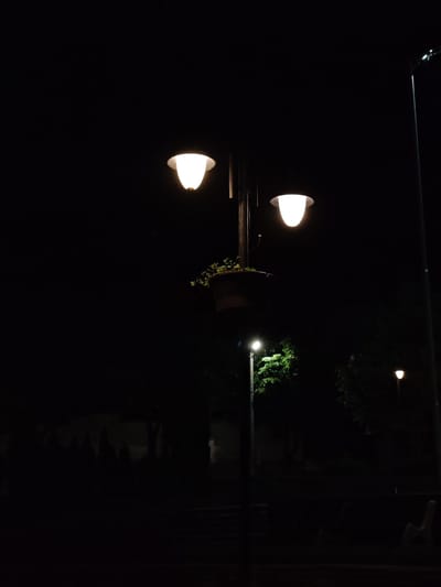 The image displays a nighttime scene in Veliki Preslav, Bulgaria, centered on a dark streetlamp pole equipped with two illuminated, bell-shaped light fixtures emitting a warm white glow. Midway down the pole, a dark planter holds green foliage. To the lower left, a second illuminated streetlamp is visible, casting light onto dark green foliage. A third, more distant, faintly illuminated streetlamp is discernible in the lower right background. The environment consists of dark trees and indistinct ground features, suggesting an outdoor public space. No individuals or animals are present.