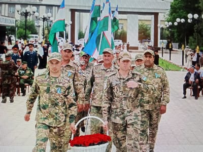 A group of men in military uniforms are walking in a procession, some holding flags and one carrying a basket of red roses. The men are wearing camouflage uniforms, and some have medals or ribbons on their chests. The flags are green and white with a crescent moon and stars, which appear to be the flag of Uzbekistan. In the background, there are buildings, streetlights, and other people watching the procession. The scene appears to be taking place in a public square or street. Based on the context of the Uzbek flag and the architecture of the buildings in the background, it is likely that this scene is taking place in Karaganda, Uzbekistan.