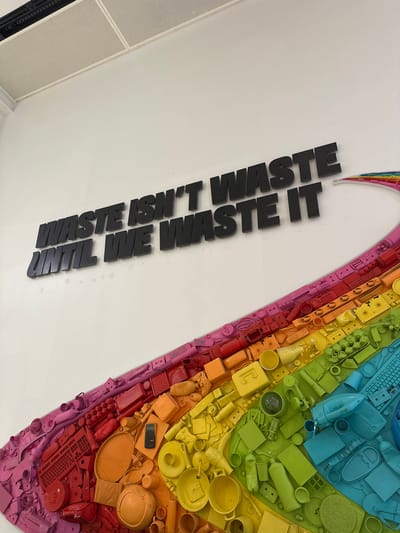 A low-angle, tilted, indoor shot shows a wall decoration consisting of the words "WASTE ISN'T WASTE UNTIL WE WASTE IT" in large, dark letters, next to a rainbow-colored heart shape made of recycled objects. The background is a bright white wall. The wall is at the office of a company called "Werfzeven" located at Tractieweg 77 in Utrecht, Netherlands.