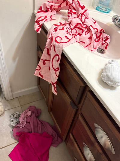 A cluttered bathroom scene shows a sink with a pink and red patterned scarf draped inside. A small, white, rolled-up towel sits next to the sink. Below, dark wood cabinets with silver handles are partially visible. More pink clothing is piled on the tiled floor next to the cabinets, near the white wall and baseboard. The photograph is taken from an elevated perspective.