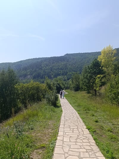 Two people walk along a paved pathway in Solnechnaya Polyana, Russia. The pathway is made of irregular light-colored stones and curves to the left as it ascends a gentle grassy slope. To the left of the path, tall green grass and wildflowers grow. To the right of the path, the grassy slope continues upwards, bordered by trees and a densely forested hillside that rises to a distant, hazy mountain range under a clear blue sky. The trees closest to the path are a mix of deciduous and coniferous species. The two individuals are positioned in the distance on the pathway, facing away from the camera and walking towards the left. The person in front wears a light-colored top and pants, while the person behind them wears a dark top and light-colored pants. The scene depicts a recreational outdoor setting on a sunny day.