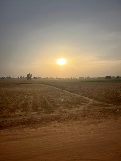 A landscape under a hazy sky, illuminated by the sun. The foreground features a field of dry grass or dirt. A line of trees or vegetation runs along the horizon. The sun appears as a bright, circular orb amidst the hazy atmosphere, casting a soft, warm light across the scene. The sky fades from light grey to a warmer tone near the sun. The image has an overall soft, muted quality.