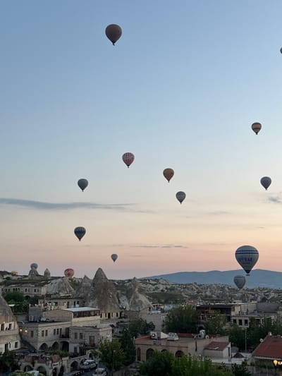 This media file captures a picturesque scene of hot air balloons soaring over a unique landscape at dawn or dusk in Göreme, Turkey.

The sky, occupying the upper and left portions of the image, displays a gradient of colors, transitioning from a clear light blue on the left to soft pink, orange, and pale yellow hues towards the right, indicating either sunrise or sunset. Wispy clouds are visible in the warmer-toned section of the sky.

Numerous hot air balloons are scattered across the sky at varying altitudes. They come in different patterns and colors; some are striped (e.g., yellow and brown, red and white), while others are solid. One prominent blue and white balloon in the mid-right clearly displays the text "ROYAL BALLOON." Another balloon, partially visible, has "DOYAL" written on it.

Below and to the right of the sky, the distinctive geological formations known as fairy chimneys are visible, characteristic of Cappadocia. These grey, conical rock structures are interspersed with a settlement of buildings. The buildings appear to be constructed from light-colored stone, some seemingly carved into the rock, with red-tiled roofs and multiple windows, suggesting a traditional architectural style. There are also some trees and other greenery among the structures.

The overall scene depicts the popular hot air balloon activity over the unique landscape of Göreme, with a sign bearing the text "KAYA" partially visible on one of the buildings on the far right. The soft lighting contributes to a serene and captivating atmosphere.