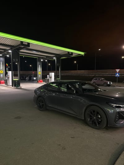 The image depicts a nighttime scene at a Toheko gas station in Ivashevo, Russia. A dark gray Volkswagen car is parked next to pump number 2. In the distance, other vehicles and individuals are visible. The gas station is well-lit with fluorescent green accents, and signage is prominent. The surrounding environment appears to be paved.