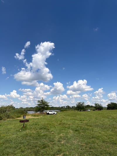 This is a horizontal, eye-level, outdoor shot of a grassy field with a bright blue sky. The sky is filled with fluffy white clouds. In the background, there is a body of water with trees and vegetation on the far bank. A white car is parked on the grassy area near the water. There is also a metal barbecue grill in the foreground. The sun is shining, indicating it is daytime. The scene appears to be a park or a recreational area. There is no visible text.