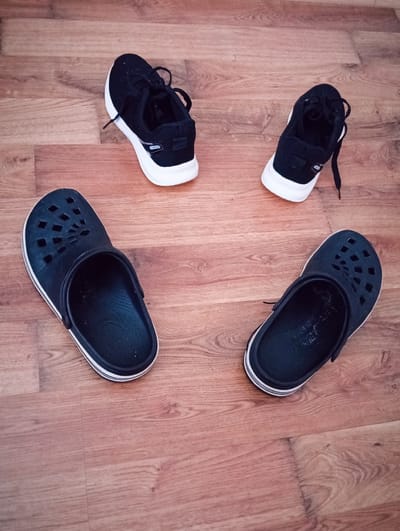 Four shoes are laid out on a wooden floor. Two black clogs with white trim are positioned on the left and right sides of the image, facing inwards. To the upper left and upper right are two black athletic shoes with white soles and laces. One of the athletic shoes has the word "FASHION" visible on its side. The scene is an indoor setting, likely a residence, with no indication of the city of Tišnov, Czechia.