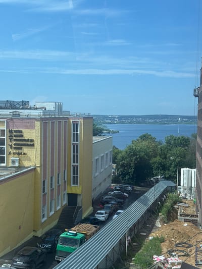 The image captures a view from an elevated position, looking towards a body of water and a cityscape in the distance. In the foreground, a yellow and pink building with signs in Russian indicating various facilities such as a swimming pool, gym, and sports hall, is visible. Several cars are parked outside this building. To the right, a construction site is in progress, with heavy machinery, piles of dirt, and building materials. A green truck loaded with sand is also present. The background features a wide river or lake, with a town or city on the opposite shore, under a clear blue sky with scattered clouds. The overall scene suggests an urban environment with ongoing development.