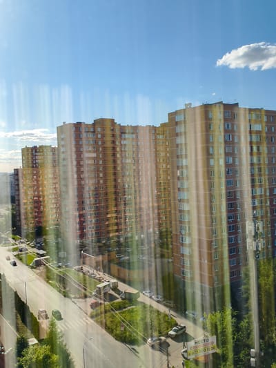 The image shows a view of a residential area in the city of Vidnoye, Russia. The primary subjects are several tall, multi-story apartment buildings with a predominantly reddish-brown and yellow facade, characterized by numerous windows. The buildings are positioned in a row, suggesting a large housing complex. In the foreground, a street runs alongside the buildings, with several cars parked and driving on it. Green spaces with trees and grass are visible between the street and the buildings, as well as surrounding the area. The sky above is blue with scattered white clouds, indicating fair weather. Vertical light streaks are present across the image, possibly due to reflections on a window. A billboard with Russian text and a phone number is visible near the street. The scene depicts a typical urban residential environment on a sunny day.