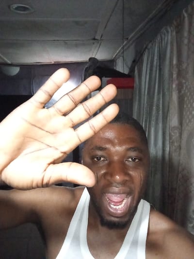 A man poses close to the camera. He's dark-skinned and wears a white tank top. He has a short beard and is grinning with his mouth open. His right hand is held up, fingers spread, partially obscuring the view. The background appears to be indoors, featuring a ceiling with a light fixture, a red object on the wall, and patterned curtains. The lighting in the room appears somewhat dim and the man looks shiny.