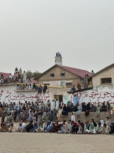 In the city of Chaman, Tehsil, Pakistan, a large gathering of men and boys is observed in front of a weathered building. The crowd is seated on the ground and on a raised concrete structure. Many individuals are wearing traditional Pakistani attire, including shalwar kameez. Some are observing a structure in the background, which appears to be a building with a red-tiled roof and a prominent clock tower-like feature. The building's facade is adorned with large red Urdu script lettering. People are also perched on the roof of the building, overlooking the scene. The overall atmosphere suggests a public event or gathering.