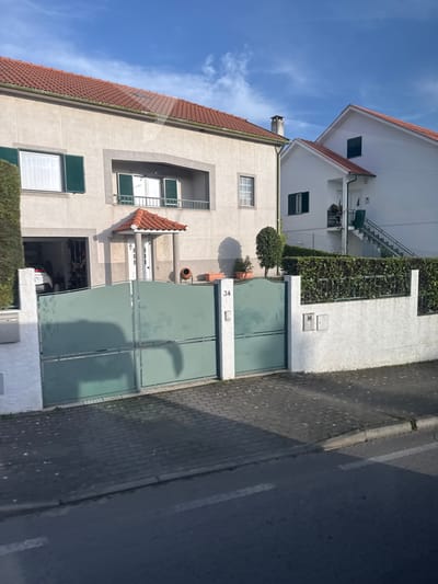 An exterior daytime photograph captures a street scene featuring two residential houses in Macedo de Cavaleiros, Portugal. The prominent house on the left is a two-story structure with a light beige stucco façade and a red terracotta-tiled roof, from which a brick chimney emerges on the right side. The ground floor includes an open garage on the far left, revealing the front portion of a white vehicle, and a central main entrance with white double doors sheltered by a small, terracotta-tiled porch roof supported by two columns. The second floor features a large window with open dark green louvered shutters on the left, a recessed central balcony with white French doors and closed dark green shutters, and a smaller window with white frames on the right. A manicured green hedge stands tall on the left, bordering the property. Near the main entrance, a small, topiary-style tree and several potted plants are visible.
To the right of the primary house, a second, white-stuccoed residence with a steeply pitched red terracotta-tiled roof is present. This house features an external metal staircase leading to an upper-level entrance and at least two windows, one of which has closed dark green shutters.
The foreground displays a large, closed, double-leaf gate in a sage green color, horizontally segmented, set between two white stucco pillars. The right pillar is marked with the number "34" and contains an intercom unit. Extending to the right from this pillar is a low white stucco wall topped with a dense, green hedge, which also features two utility boxes. The property is separated from the street by a sidewalk paved with dark grey interlocking bricks. An asphalt street with white dashed lines occupies the immediate foreground. Bright sunlight from the left casts long shadows from the gate and houses across the sidewalk and street. No individuals are present in the scene.