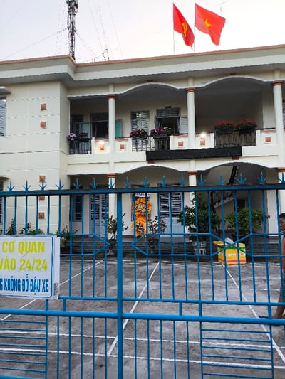 The image depicts a two-story building in Thành phố Quảng Ngãi, Vietnam, viewed from outside a blue metal gate. The building is light-colored, likely cream, with brown accents on columns and trim. Two red flags fly from the rooftop: the flag of the Communist Party of Vietnam (yellow hammer and sickle) and the national flag of Vietnam (yellow star). Above the building, a metal communication tower is visible against an overcast sky.
The first floor features a long balcony with potted plants containing red, purple, and white flowers, situated in black rectangular planters. Windows with light-colored frames and shutters are open. The ground floor exhibits arched doorways or openings, with additional potted plants arranged in the foreground. A prominent orange and yellow floral arrangement stands near a central entrance.
A blue metal gate, composed of vertical and horizontal bars forming rectangular and arrow-shaped patterns, spans the foreground. Attached to the left side of the gate is a white rectangular sign with yellow text: "CƠ QUAN VÀO 24/24" (Agency/Office accessible 24/7) and "KHÔNG ĐỖ ĐẦU XE" (No parking).
Behind the gate, a paved courtyard surface features white line markings. On the right side of the courtyard, a multi-colored box (yellow, pink, blue, white) rests near a visible outdoor staircase. A person's lower body, wearing light-colored trousers and sandals, is partially visible on the far right edge of the frame, walking to the left.