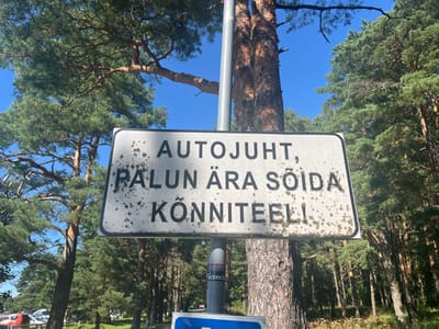 A sign in Estonian reads "AUTOJUHT, PALUN ÄRA SÕIDA KÕNNITEELI," which translates to "DRIVER, PLEASE DO NOT DRIVE ON THE SIDEWALK." The sign is white with black lettering and is mounted on a metal pole. It is surrounded by trees with green foliage and a bright blue sky. The sign appears to be old and weathered, with some dirt and lichen on its surface. In the background, there is a glimpse of cars parked in a lot, suggesting an urban or semi-urban environment. The overall scene suggests a public area where pedestrian safety is being emphasized.