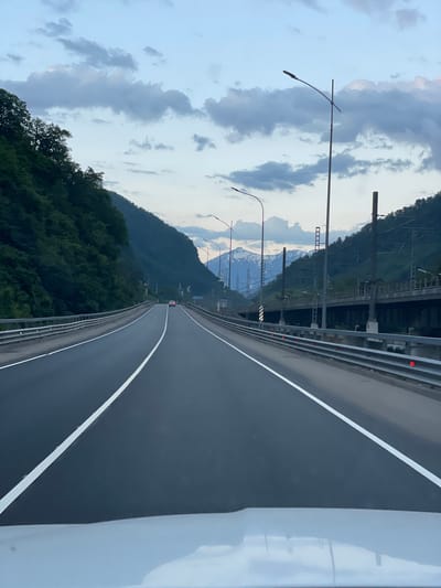 The image captures a view from a vehicle driving on a paved road in Chvizhepse, Russia. The road is divided by white lines, indicating multiple lanes. On the left side of the road, a steep, tree-covered mountainside rises. On the right, a metal guardrail runs along the edge of the road, and beyond that, an elevated structure, possibly a bridge or viaduct with train tracks, is visible. In the distance, snow-capped mountains are visible under a cloudy sky. A single white car is seen driving ahead on the road. Streetlights are positioned along the road.