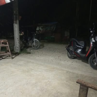 The image depicts a dimly lit outdoor scene in Matangmeunye, Indonesia at night. Two motorcycles are parked on a concrete and gravel surface. One is a dark scooter with red accents, parked on the right side of the image. The other is a lighter-colored, older model motorcycle parked further back, near what appears to be a wooden structure with a tattered cover. A wooden stepladder stands to the left of a concrete pillar. A partial view of a wooden structure is also visible in the lower right corner.