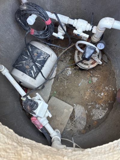 The image shows a round, concrete pit in Uyutnoye, Ukraine, containing plumbing equipment. In the center of the pit is a white pressure tank with a black metal bracket on top. To the right of the tank is a circular component with a pressure gauge attached to a white pipe. Multiple white PVC pipes and a black electrical cable are visible throughout the pit, some secured with red tape. The pit floor is covered in dirt, small rocks, and debris.
