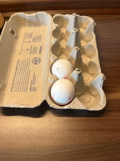 The image shows two white eggs in a cardboard egg carton. The carton is open and the two eggs are in the last two slots. The eggs have pink markings. The carton is sitting on a wooden surface. The eggs appear to be in the city of Köln, Germany, located at latitude 50.934, longitude 6.974. The carton has writing in German on the bottom portion, which is partially visible in the image. The writing includes the word "Frische", which means "fresh". The writing indicates the eggs are from Germany and were produced in accordance with German quality standards.  The eggs are not being interacted with. The wooden surface is light brown in color.