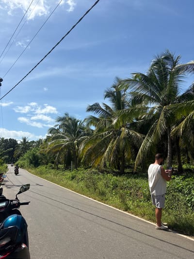 A man stands on the side of a paved road, holding a cell phone. He is wearing a sleeveless white t-shirt and light gray shorts. He is also wearing white slip-on shoes. He is standing in front of a row of palm trees. The road is wet and there are two motorbikes on the side of the road. The sky is blue with a few white clouds. The location is Unknown, Sri Lanka.