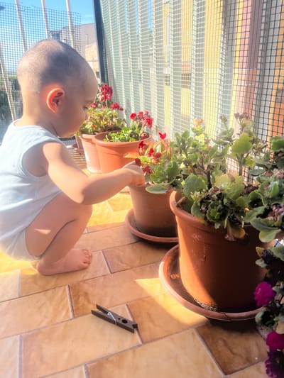 The image shows a young child crouching on a tiled balcony in Stornara, Italy, interacting with potted plants. The child, wearing a light blue tank top and diaper, is reaching towards a plant in a terracotta pot, appearing to touch its leaves. In the foreground, a wooden clothespin lies on the tiled floor. Behind the child and plants, a white mesh netting covers the balcony railing, and beyond that, a yellow wall and buildings are visible under a bright sky. The scene is bathed in warm sunlight, casting shadows on the tiles.