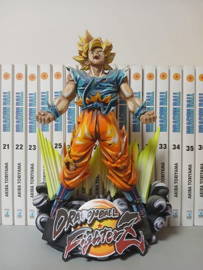 The image shows a figurine of Goku from Dragon Ball Z standing in front of a row of Dragon Ball manga volumes. The figurine is in a dynamic pose, with its arms outstretched and its mouth open in a shout. Goku is wearing his iconic orange gi, which is ripped in places, revealing his blue undershirt. The manga volumes are white with the Dragon Ball logo and the author's name, Akira Toriyama, printed on them. The spines of the books are numbered sequentially, starting from 21 and going up to 36. The figurine is placed on a circular base with the "Dragon Ball FighterZ" logo. The background is a plain white wall. The image appears to be taken indoors, possibly in a home or a store. There is no indication of the location being in Monsummano Terme, Italy, or any other specific city.