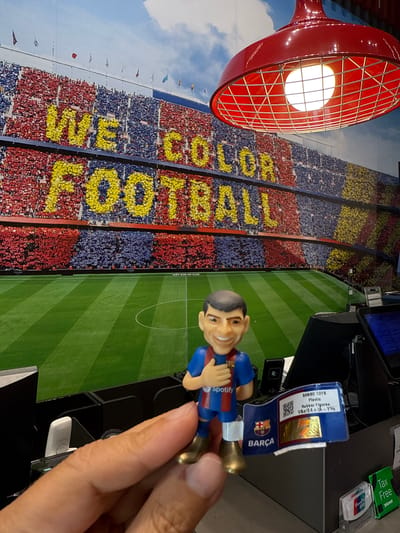 A person's right hand is holding a small, caricature-like figurine of a soccer player wearing a blue and red FC Barcelona jersey with a Spotify logo. The figurine has gold-colored shoes and a medal around its neck. The figurine is standing in front of a backdrop depicting a packed soccer stadium with the words "WE COLOR FOOTBALL" written in yellow letters across the stands. In the foreground, on a counter, are various electronic devices and a price tag for the figurine, which reads "BANBO TOYS Plastic Rubber Figures". A red hanging lamp with a caged bulb is visible in the upper right portion of the image. The scene is likely within a retail environment, possibly a store at La Roca Village in La Roca del Vallès, Spain, given the backdrop and the presence of tax-free shopping information.