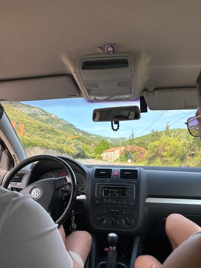 The image provides an interior perspective from a Volkswagen car, looking forward through the windshield at a scenic, mountainous landscape in Donji Katun, Montenegro.

In the foreground, the driver is partially visible on the left, wearing a white t-shirt, with hands on a black three-spoke steering wheel featuring the Volkswagen logo. Keys hang from the ignition. The dashboard displays a speedometer, tachometer, and a digital readout showing "17:39" and "7.2". The center console includes air vents, an infotainment system displaying "Info", "Песня без сл" (Song without words), and "OB", below which are climate controls and a 5-speed manual gear shifter.

To the right, a passenger's upper arm and legs are partially visible. Sunglasses are resting on the passenger's head, out of frame. The beige car ceiling features an overhead console, sun visors, and a small round pin depicting a person's face attached to the liner near the driver's side. A black beaded ornament hangs from the rearview mirror. A small dashcam or similar device is mounted to the right of the rearview mirror, and a red and white striped ribbon is attached to the ceiling liner.

Beyond the windshield, a paved road curves through a predominantly green, mountainous environment. The mountainsides are covered with lush vegetation, including trees and dense bushes. A stone wall lines the immediate roadside. In the distance, traditional-style houses with red-tiled roofs are nestled among the foliage, with one prominent house having vibrant pink flowering bushes nearby. Construction activity is evident, with orange scaffolding or machinery visible on the left mountainside and a yellow crane further back on the right. Power lines traverse the clear blue sky, which shows scattered white clouds. The overall scene depicts a journey through a picturesque, hilly region.