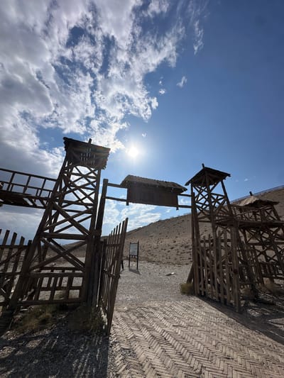 The image shows a rustic wooden gateway structure with two guard towers, likely part of a historical reenactment or themed attraction in Jiayuguan, China. The sun is bright, casting strong shadows. The gateway is open, leading to a gravel path and a barren, hilly landscape. The architecture of the wooden structures appears old and weathered, suggesting a frontier or historical setting. The sky is partly cloudy with the sun visible. In the background, a gentle slope is covered with dry vegetation and rocks. A signpost is visible to the right of the gateway, and a person can be seen walking away from the camera in the distance, also to the right. The ground in the foreground is paved with a herringbone brick pattern. The overall impression is one of a dry, arid environment with a focus on historical reconstruction.