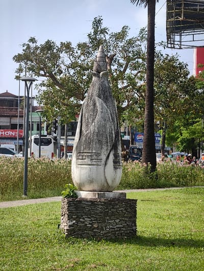 The image depicts an outdoor urban scene in Thuận Hóa Ward, Vietnam, under clear daylight conditions.
In the foreground, a tall, weathered, light-colored stone sculpture stands centrally on a rectangular base constructed from stacked, dark, irregular stones. The sculpture features an elongated, abstract, bulbous form, tapering to a multi-tiered, conical finial at its apex. Its surfaces exhibit significant grime and discoloration. Carved relief patterns, including horizontal lines and geometric shapes, are visible on its lower half. Small green plants are growing from the top edge of the stone base on the left side. The sculpture rests on a manicured green lawn, which casts a shadow to the right.
Behind the sculpture, a band of tall, wispy, light-brown grasses or reeds separates the lawn from a paved roadway. A dark-grey, multi-headed street lamp with a curving arm is positioned to the left, rising from these grasses.
The midground and background reveal a bustling street environment. Numerous mature trees with dense green foliage provide a natural backdrop, including a slender, dark-barked palm tree trunk on the right. Beyond the trees, multi-story urban buildings line the street, displaying typical Vietnamese architecture with balconies and storefronts. Visible signage includes "HON" and "NHON" on the left, and a "PEPSI" logo partially visible on the right. Traffic is present on the road, with a white passenger van (bearing "403.11") moving from left to right, and several motorbikes and scooters with riders further down the road to the right. A large metal scaffolding structure or billboard frame is partially visible above the trees on the far right. The sky overhead is clear and light blue.
