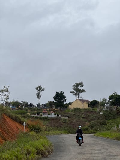 The scene depicts a motorcyclist traveling on a paved road in Trường Thọ, Vietnam. The rider wears a helmet and dark clothing, with bright red footwear. The road is lined with dense vegetation on the right and a terraced landscape with various structures and trees on the left. In the background are residential buildings and a predominantly overcast sky. The foreground includes a reddish-brown soil embankment.