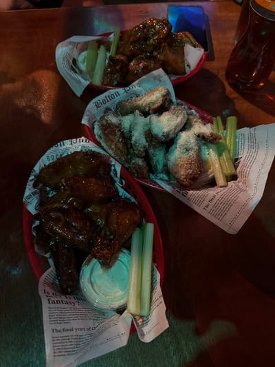 The image depicts a table with three baskets of chicken wings, celery sticks, and a small container of sauce. Each basket is lined with newspaper print. A brown bottle is visible at the top left, and a cell phone is in the upper left corner. The image appears to have been taken in a restaurant or bar setting, likely in Tuxtla Gutiérrez, Mexico, based on the OCR text on the newspaper.