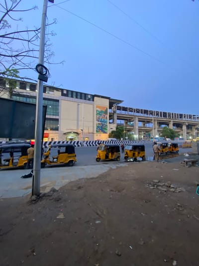 The image depicts a street scene in St. Thomas Mount-Pallavaram Cantonment Board, India, likely during the late afternoon or early evening due to the soft lighting and clear sky. The primary subjects are several yellow auto-rickshaws parked along the side of a road. A few individuals, some appearing to be drivers or pedestrians, are visible near the vehicles.

The scene is set against the backdrop of a multi-story building with a light-colored facade and several floors of windows. A pedestrian walkway with a black and white striped barrier runs alongside the road. To the right of the walkway, the ground is uneven and appears to be a dirt or gravel area with scattered debris. Overhead, power lines stretch across the frame, and a metal structure, possibly part of an elevated road or construction, is visible in the background.

A large mural or advertisement featuring colorful imagery is painted on the side of one of the buildings. The overall atmosphere is that of a typical urban street in India, with the presence of auto-rickshaws suggesting a mode of public transportation. There is no discernible text visible in the image. The lighting suggests it's not midday, with the sky being a clear, light blue.